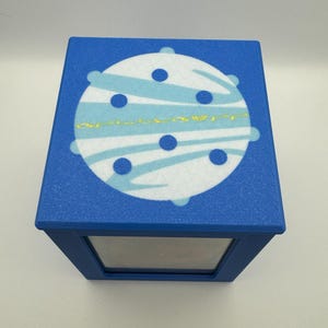 May include: Blue box with a white and blue planet design on the lid. The planet has blue stripes and dark blue dots. The box has a mirrored front.