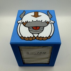 May include: A blue card storage box featuring a cartoon illustration of a white bison with brown horns and a brown arrow on its head. The box has the text "3D.ISH DESIGNS" on the front.