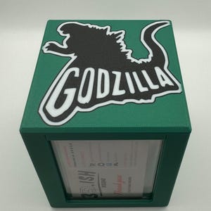 May include: A green box with a Godzilla graphic on top. The Godzilla image is black with a white outline and the word "GODZILLA" in white. The box has a clear window on the front.