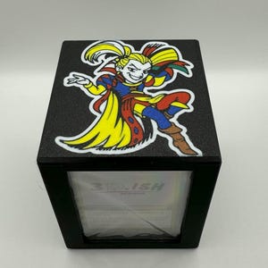 May include: Black box with a colorful cartoon image of a character on top. The character has yellow hair, a yellow cape, and is wearing blue, red, and brown clothing. The text "3RD.ISH DESIGNS" is visible on the front.