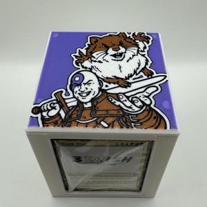 May include: A white card storage box with a purple lid featuring a cartoon illustration. The lid has a smiling bald character with a sword and a brown cartoon animal. The box has the text "Thank You!" and "3DISH".