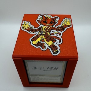 May include: A red card storage box with a cartoon character patch on the lid. The patch depicts a red demon-like figure with yellow flames in its hands. The box has a card visible with the text "3D-ISH DESIGNS".