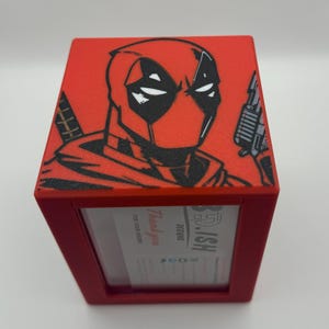 May include: Red napkin holder featuring a black and white illustration of Deadpool. The top of the holder has the character's face and a gun. The front of the holder has a clear window and text that reads "Thank you for your order."