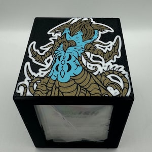 May include: Black tissue box with a detailed illustration on the top. The artwork features a blue and gold creature with intricate details and a white outline. The tissue box has a rectangular shape and a visible opening for tissue dispensing.