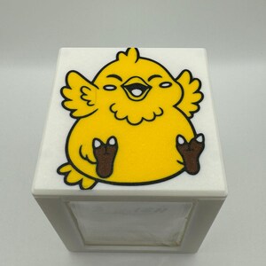 May include: A white tissue box with a smiling yellow chick illustration on top. The chick has brown feet and a black outline. The tissue box is a square shape and has a slot for dispensing tissues.