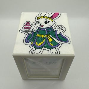 May include: White, cube-shaped tissue box featuring a cartoon rabbit wearing a green and purple outfit with a yellow sash and a flower crown. The rabbit holds a tray of pink pastries. The tissue box has the word "SO...ISH" printed on the front.