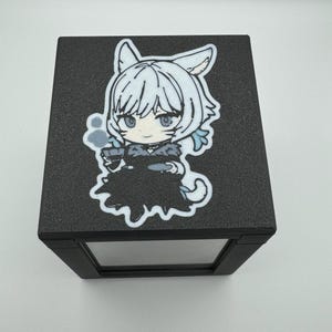 May include: A black display box with a clear front panel. The top of the box features a colorful cartoon graphic of a character with white hair, cat ears, and a black dress. The character is holding a small cup.