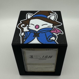 May include: Black card storage box featuring a cartoon character with a brown hat and blue cape. The box has a clear window displaying a card with the text "3D.ISH DESIGNS". The top of the box has a textured black surface.