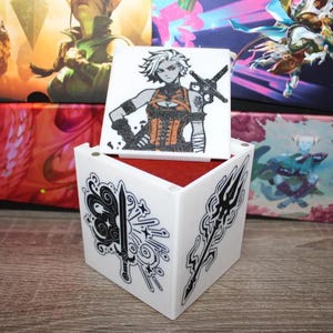 May include: A white, cube-shaped box with black and white graphic designs of weapons and symbols. A square lid features a character illustration. The box is open, revealing a red interior. Other boxes with fantasy art are in the background.