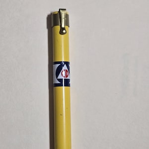 May include: A yellow metal pen with a gold-colored clip. The pen has a white and blue emblem with the letters "CD" on it. The pen is cylindrical and appears to be vintage.