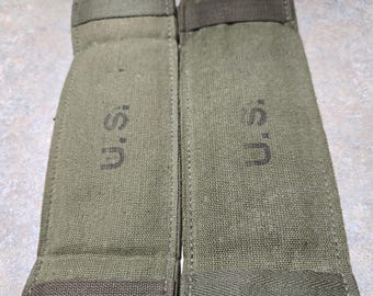May include: Two olive drab canvas pouches with the letters "U.S." printed on them. Each pouch has a rectangular body with a flap at the top and a strap. The pouches are likely military surplus or vintage items.