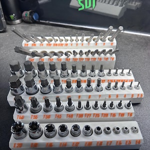 May include: A set of various metal socket bits and hex bits, organized in white plastic trays. The bits are of different sizes and shapes, including star, hex, and torx. The trays are labeled with size markings. The background is a dark surface.