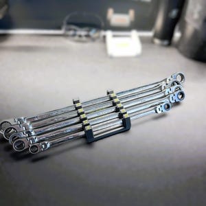 Puede incluir: Un juego de seis llaves combinadas plateadas con mangos negros y marcas amarillas. Las llaves están dispuestas en un soporte negro con los números 8, 10, 12, 14, 15 y 17 impresos en él.