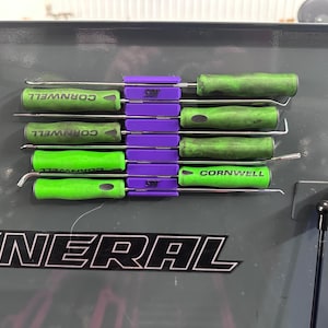 May include: A set of six green handled tools with silver metal tips are held in place by a purple magnetic strip. The tools are labelled "Cornwell" and are mounted on a grey metal surface with the word "General" printed in black.