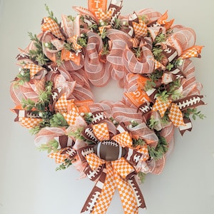 May include: A decorative wreath featuring a football theme. The wreath is made of orange, white, and brown mesh, with football-shaped accents and ribbons. Greenery and small white accents are interspersed throughout the design. The wreath is circular.