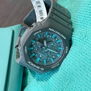 Puede incluir: Un reloj G-Shock gris con una esfera azul y blanca. El reloj tiene una pantalla digital y una variedad de botones, incluyendo "Start", "Mode", "Light" y "Adjust".