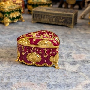 May include: A heart-shaped trinket box with a red base and gold accents. The box is decorated with intricate floral patterns and has a hinged lid.