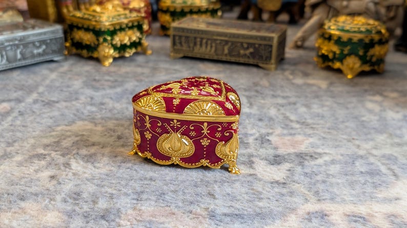 May include: A heart-shaped trinket box with a red base and gold accents. The box is decorated with intricate floral patterns and has a hinged lid.