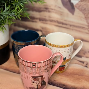 May include: Three ceramic mugs with Egyptian-themed designs. The front mug is pink with hieroglyphs and a pharaoh's portrait. The other mugs are blue and white, also with Egyptian motifs. The mugs have gold accents and white handles. A green plant is in the background.