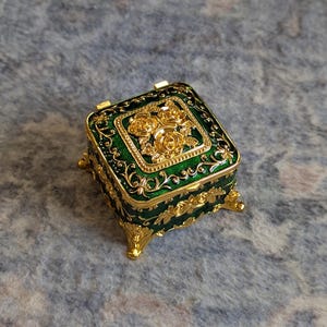 May include: A small, square, green and gold trinket box with ornate floral designs. The box has four feet and a hinged lid.