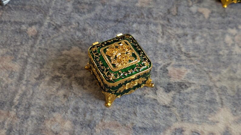 May include: A small, square, green and gold trinket box with ornate floral designs. The box has four feet and a hinged lid.