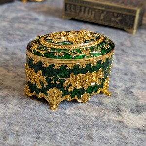 May include: A small, oval-shaped trinket box with a green enamel finish and gold floral designs. The box has four gold feet and a decorative gold trim.