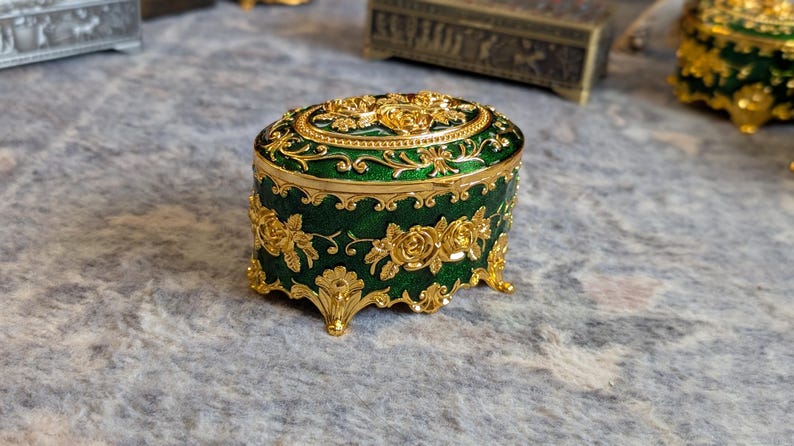 May include: A small, oval-shaped trinket box with a green enamel finish and gold floral designs. The box has four gold feet and a decorative gold trim.