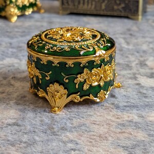 May include: A small, round, green trinket box with gold accents and floral designs. The box has four legs and a hinged lid.