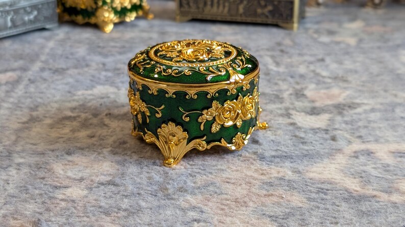 May include: A small, round, green trinket box with gold accents and floral designs. The box has four legs and a hinged lid.