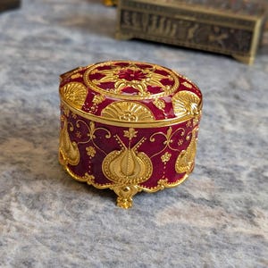 May include: A small, round, red and gold trinket box with intricate floral designs. The box has a lid and sits on four small feet.