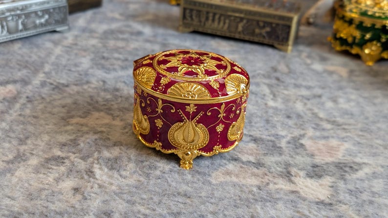 May include: A small, round, red and gold trinket box with intricate floral designs. The box has a lid and sits on four small feet.