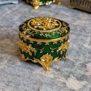 May include: A small, round, green trinket box with gold accents and floral designs. The box has four feet and a hinged lid.