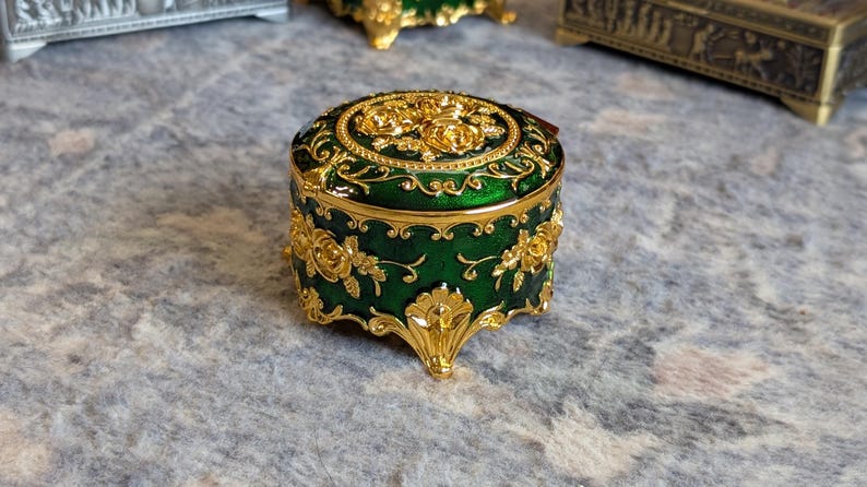 May include: A small, round, green trinket box with gold accents and floral designs. The box has four feet and a hinged lid.