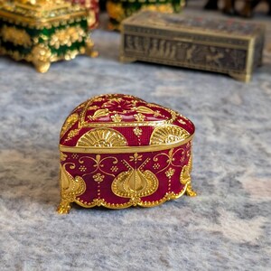 May include: A heart-shaped trinket box with a red base and gold accents. The box is decorated with intricate floral and geometric patterns.