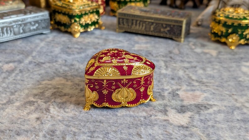 May include: A heart-shaped trinket box with a red base and gold accents. The box is decorated with intricate floral and geometric patterns.