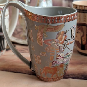 Puede incluir: Taza de cerámica gris con un asa grande, con un diseño de temática egipcia. La taza tiene una base gris claro con ilustraciones de jeroglíficos y figuras doradas y naranjas. El borde y la base tienen bordes decorativos. Otras tazas son visibles en el fondo.