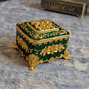 May include: A small, square, green and gold trinket box with ornate floral designs. The box has four decorative feet and a hinged lid.