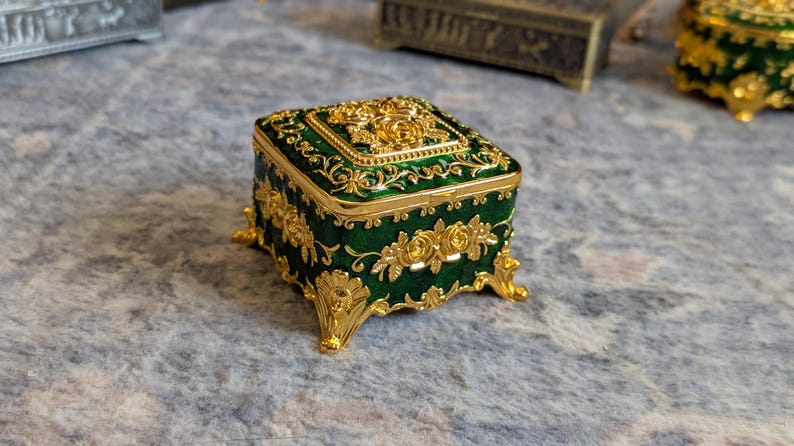 May include: A small, square, green and gold trinket box with ornate floral designs. The box has four decorative feet and a hinged lid.