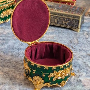 May include: A small, round, green and gold trinket box with a red velvet interior. The box has a floral design and sits on four gold feet.