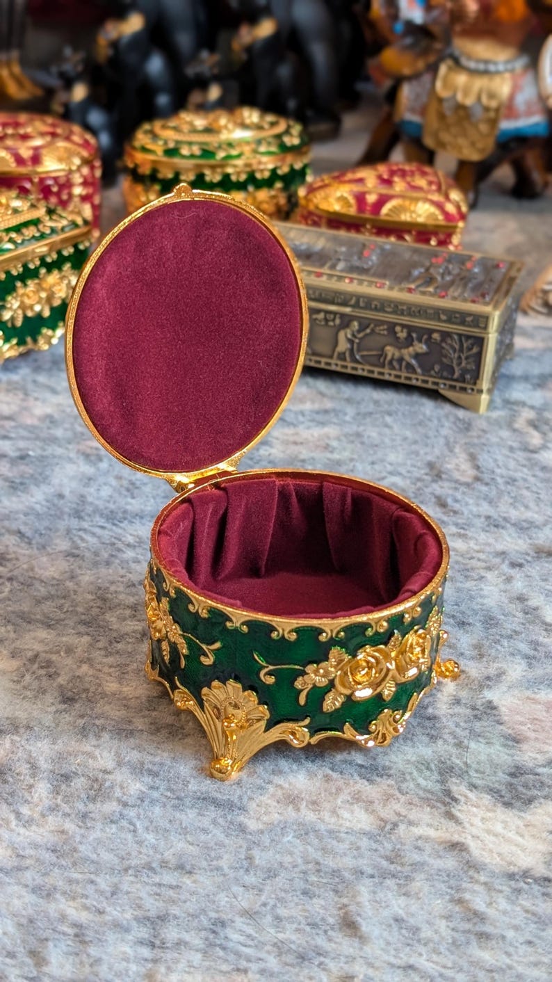 May include: A small, round, green and gold trinket box with a red velvet interior. The box has a floral design and sits on four gold feet.