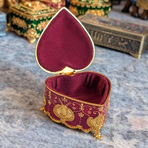 May include: A heart-shaped trinket box with a red velvet interior and gold accents. The box is decorated with a floral pattern and has a hinged lid.