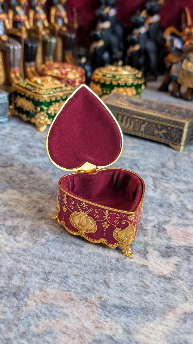 May include: A heart-shaped trinket box with a red velvet interior and gold accents. The box is decorated with a floral pattern and has a hinged lid.