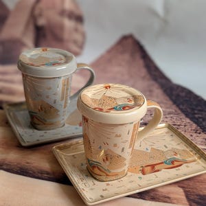 May include: Two ceramic mugs with lids and rectangular trays. The mugs and trays feature an Egyptian design with pyramids, hieroglyphs, and gold accents. The mugs have handles and are light beige and grey.