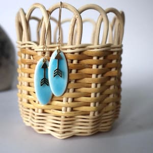 May include: A pair of blue ceramic earrings with gold arrow charms hanging from a wicker basket.