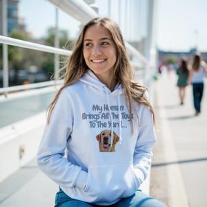 May include: A white hooded sweatshirt featuring a graphic of a golden retriever dog and the text "My Human Brings All The Toys To The Yard..." printed in dark grey lettering on the front chest area, complete with a front kangaroo pocket.