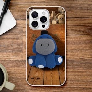 May include: A smartphone case featuring a graphic of a blue and gray donkey character with a black tuft of hair on its head. The character has a rounded gray face, blue ears, and white patches on its feet, set against a wooden background.