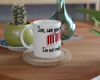 May include: A white ceramic mug featuring black text that reads, Son, can you play, and I'm not really, with a graphic of red and white piano keys positioned between the two lines of text. The mug has a standard handle and a cylindrical shape.