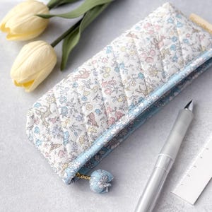 以下が含まれることがあります： パステルカラーの花柄のキルティングペンケース。小さな鳥やウサギが描かれています。ケースは水色のトリムとマッチするタッセルが付いています。銀色のペンと定規がケースの隣にあります。
