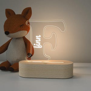 May include: A wooden night light with a clear acrylic letter 'F' that has the name 'kinn' written on it. The light is on and glowing.