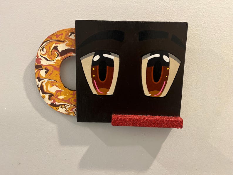 May include: A quirky wall art piece in the shape of a cartoon face. The black face has large eyes with brown irises and a red, glittery mouth. The handle is a circular shape with a marbled pattern in orange, brown, and white. A unique decorative item.